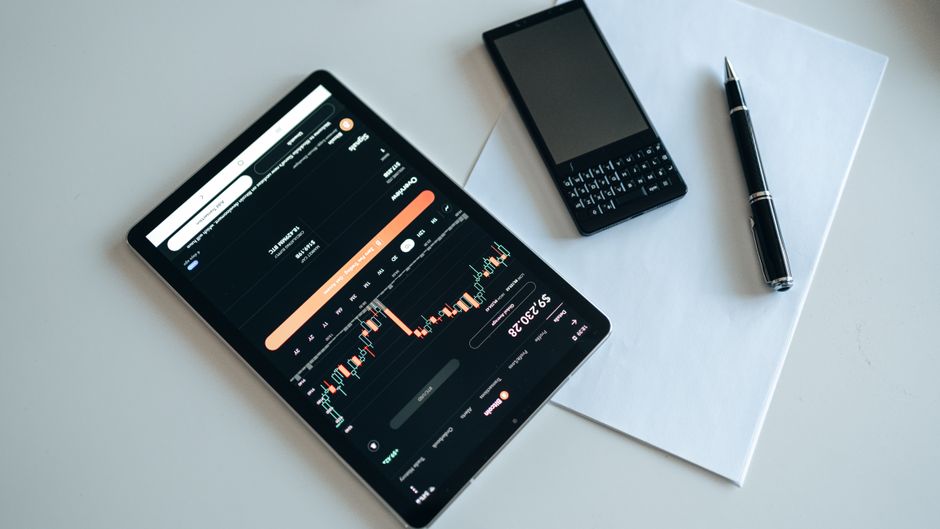 Top view of a workspace with a tablet displaying financial charts, a smartphone, pen, and paper.