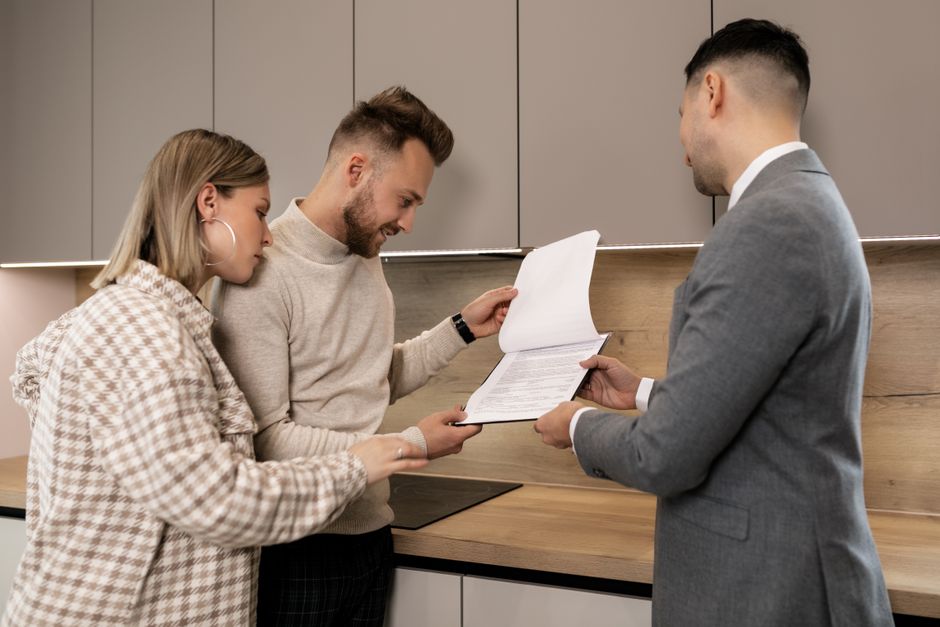 A couple reviews real estate documents with an agent in a modern indoor setting, discussing a potential property purchase.